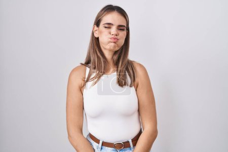 Foto de Hispanic young woman standing over white background puffing cheeks with funny face. mouth inflated with air, crazy expression. - Imagen libre de derechos