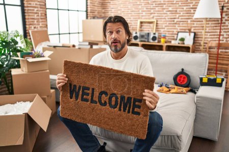 Photo for Handsome middle age man holding welcome doormat at new home in shock face, looking skeptical and sarcastic, surprised with open mouth - Royalty Free Image