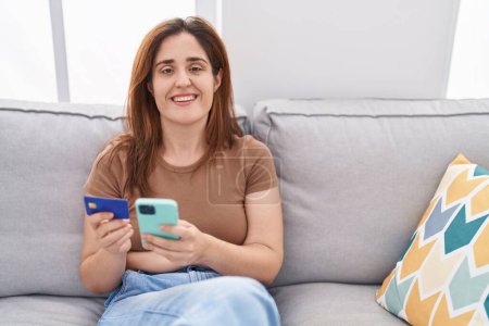 Téléchargez les photos : Young woman using smartphone and credit card sitting on sofa at home - en image libre de droit