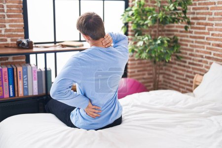 Téléchargez les photos : Jeune homme caucasien souffrant d'une blessure au dos assis sur le lit dans la chambre - en image libre de droit