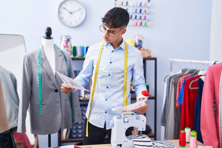 Téléchargez les photos : Jeune homme hispanique tailleur regardant la conception de vêtements au magasin de tailleur - en image libre de droit
