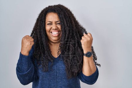 Foto de Plus size hispanic woman standing over white background very happy and excited doing winner gesture with arms raised, smiling and screaming for success. celebration concept. - Imagen libre de derechos