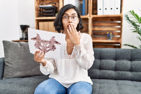 Foto de Young hispanic therapist woman holding rorschach test covering mouth with hand, shocked and afraid for mistake. surprised expression - Imagen libre de derechos