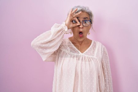 Foto de Mujer de mediana edad con el pelo gris de pie sobre el fondo rosa haciendo gesto ok conmocionado con la cara sorprendida, ojos mirando a través de los dedos. expresión incrédula. - Imagen libre de derechos