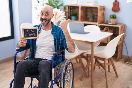 Photo for Hispanic man with beard sitting on wheelchair at new home pointing thumb up to the side smiling happy with open mouth - Royalty Free Image