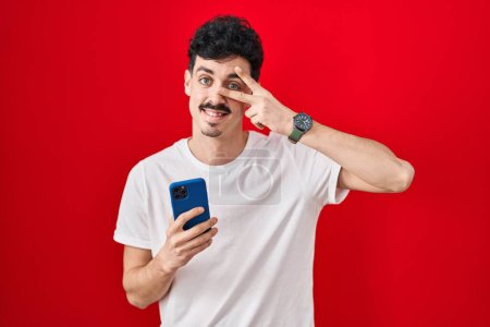 Téléchargez les photos : Hispanic man using smartphone over red background doing peace symbol with fingers over face, smiling cheerful showing victory - en image libre de droit
