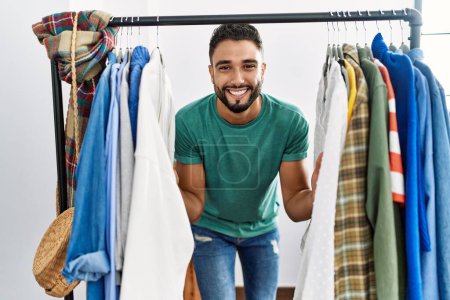 Foto de Joven árabe hombre cliente sonriendo confiado que aparece a través de perchero de ropa en tienda de ropa - Imagen libre de derechos