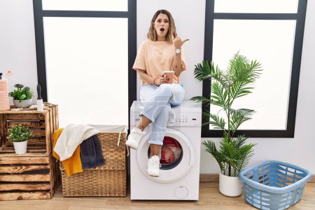 Foto de Young hispanic woman drinking coffee waiting for washing machine at laundry room surprised pointing with hand finger to the side, open mouth amazed expression. - Imagen libre de derechos