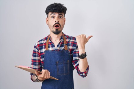Foto de Young hispanic man with beard wearing waiter apron holding clipboard surprised pointing with hand finger to the side, open mouth amazed expression. - Imagen libre de derechos