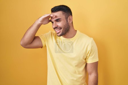 Photo for Young hispanic man standing over yellow background very happy and smiling looking far away with hand over head. searching concept. - Royalty Free Image