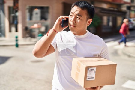 Photo for Young chinese man courier talking on the smartphone holding package at street - Royalty Free Image