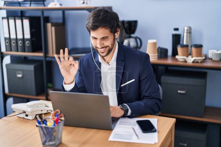 Téléchargez les photos : Bel homme latin travaillant au bureau à l'aide d'un ordinateur portable faisant signe ok avec les doigts, sourire geste amical excellent symbole - en image libre de droit