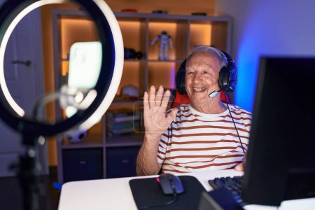 Foto de Hombre mayor con gris grabación de pelo gamer tutorial con teléfono inteligente en casa con aspecto positivo y feliz de pie y sonriendo con una sonrisa confiada mostrando los dientes - Imagen libre de derechos