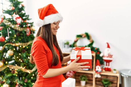 Téléchargez les photos : Jeune femme latine tenant cadeau debout par arbre de Noël à la maison - en image libre de droit