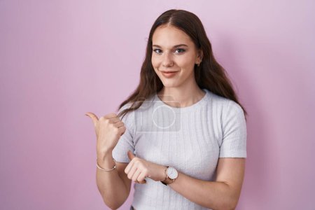 Foto de Young hispanic girl standing over pink background pointing to the back behind with hand and thumbs up, smiling confident - Imagen libre de derechos