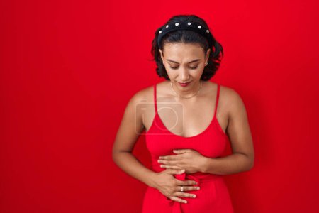 Téléchargez les photos : Young hispanic woman standing over red background with hand on stomach because indigestion, painful illness feeling unwell. ache concept. - en image libre de droit