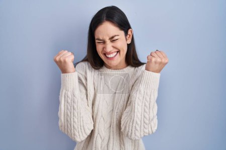 Photo for Young brunette woman standing over blue background very happy and excited doing winner gesture with arms raised, smiling and screaming for success. celebration concept. - Royalty Free Image