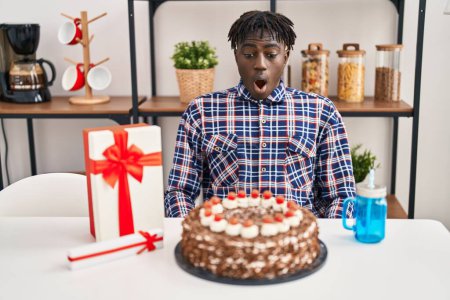 Photo for African man with dreadlocks celebrating birthday holding big chocolate cake scared and amazed with open mouth for surprise, disbelief face - Royalty Free Image