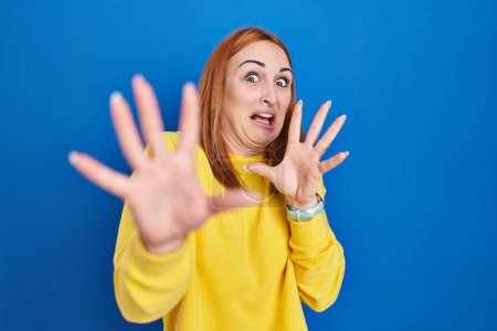 Foto de Young woman standing over blue background afraid and terrified with fear expression stop gesture with hands, shouting in shock. panic concept. - Imagen libre de derechos