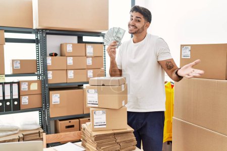 Téléchargez les photos : Young hispanic man working at small business ecommerce holding dollars celebrating achievement with happy smile and winner expression with raised hand - en image libre de droit