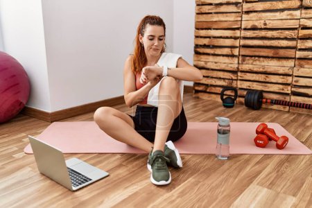 Photo for Young redhead woman watching training tutorial at the gym checking the time on wrist watch, relaxed and confident - Royalty Free Image