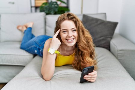 Photo for Young caucasian woman lying on the sofa using smartphone smiling pointing to head with one finger, great idea or thought, good memory - Royalty Free Image