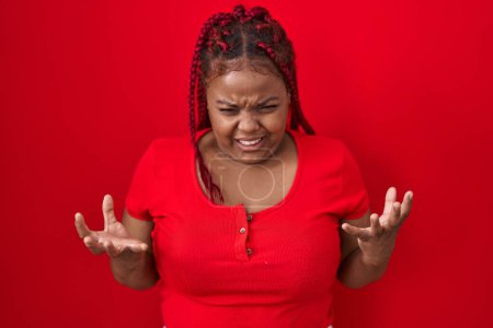 Téléchargez les photos : African american woman with braided hair standing over red background crazy and mad shouting and yelling with aggressive expression and arms raised. frustration concept. - en image libre de droit