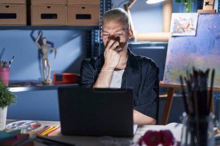 Photo for Young caucasian man using laptop at night at art studio smelling something stinky and disgusting, intolerable smell, holding breath with fingers on nose. bad smell - Royalty Free Image