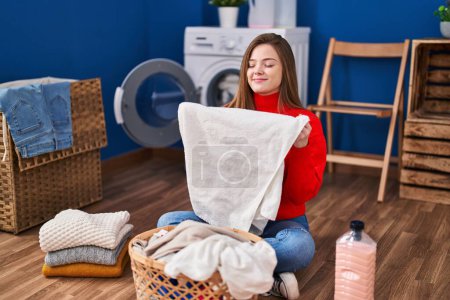 Foto de Mujer rubia joven oliendo la ropa sentada en el suelo en la lavandería - Imagen libre de derechos