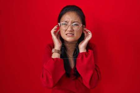 Téléchargez les photos : Asian young woman standing over red background covering ears with fingers with annoyed expression for the noise of loud music. deaf concept. - en image libre de droit