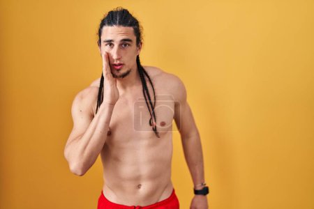 Téléchargez les photos : Hispanic man with long hair standing shirtless over yellow background hand on mouth telling secret rumor, whispering malicious talk conversation - en image libre de droit