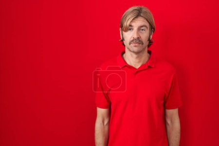 Foto de Caucasian man with mustache standing over red background relaxed with serious expression on face. simple and natural looking at the camera. - Imagen libre de derechos