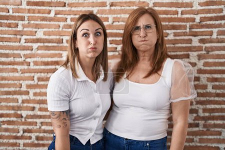 Téléchargez les photos : Hispanic mother and daughter wearing casual white t shirt puffing cheeks with funny face. mouth inflated with air, crazy expression. - en image libre de droit
