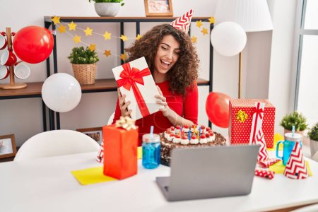 Téléchargez les photos : Young hispanic woman celebrating online birthday holding gift at home - en image libre de droit