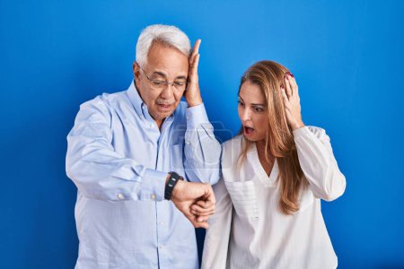 Téléchargez les photos : Couple hispanique du moyen âge debout sur fond bleu regardant le temps de la montre inquiet, peur de se faire tard - en image libre de droit
