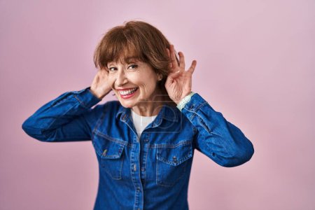 Photo for Middle age woman standing over pink background trying to hear both hands on ear gesture, curious for gossip. hearing problem, deaf - Royalty Free Image
