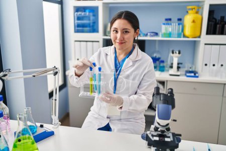 Téléchargez les photos : Young hispanic woman wearing scientist uniform holding test tubes at laboratory - en image libre de droit