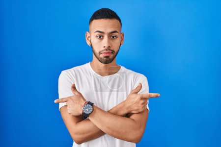Photo for Young hispanic man standing over blue background pointing to both sides with fingers, different direction disagree - Royalty Free Image