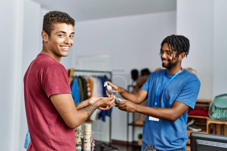 Foto de Dos hombres tendero y cliente manos de limpieza usando gel desinfectante en la tienda de ropa - Imagen libre de derechos