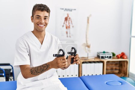 Photo for Young hispanic man working as physiotherapist holding hand grip at physiotherapy room - Royalty Free Image