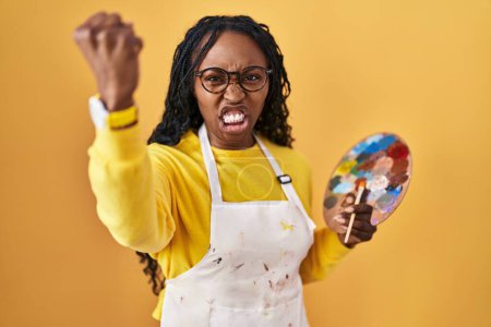 Téléchargez les photos : Femme africaine tenant palette peintre en colère et fou levant le poing frustré et furieux tout en criant de colère. rage et concept agressif. - en image libre de droit