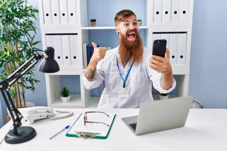 Téléchargez les photos : Rousse homme à longue barbe faire appel vidéo consulter avec smartphone pointant pouce vers le côté souriant heureux avec bouche ouverte - en image libre de droit