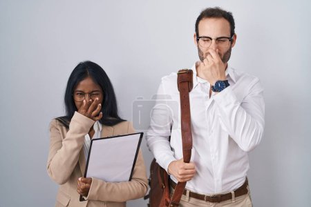 Foto de Interracial business couple wearing glasses smelling something stinky and disgusting, intolerable smell, holding breath with fingers on nose. bad smell - Imagen libre de derechos