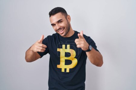 Foto de Young hispanic man wearing bitcoin t shirt pointing fingers to camera with happy and funny face. good energy and vibes. - Imagen libre de derechos