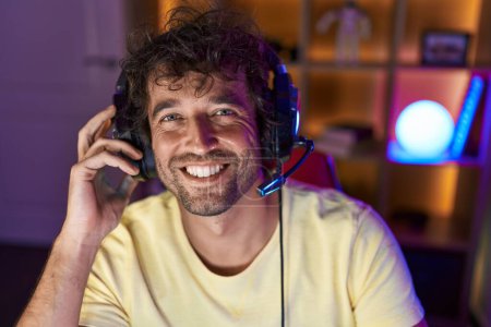 Téléchargez les photos : Jeune homme hispanique banderole souriant confiant assis sur la table à la salle de jeux - en image libre de droit