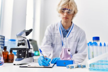 Téléchargez les photos : Jeune homme blond scientifique écrit sur un document pesant une éprouvette au laboratoire - en image libre de droit