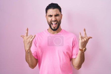 Foto de Joven hispano parado sobre fondo rosa gritando con expresión loca haciendo símbolo de roca con las manos arriba. Estrella de la música. concepto de música pesada. - Imagen libre de derechos