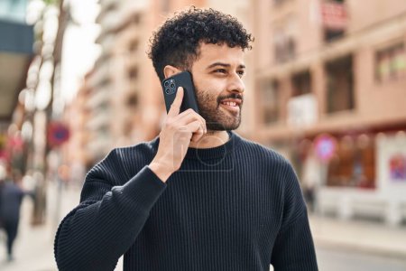 Téléchargez les photos : Jeune homme arabe souriant confiant parler sur le smartphone à la rue - en image libre de droit