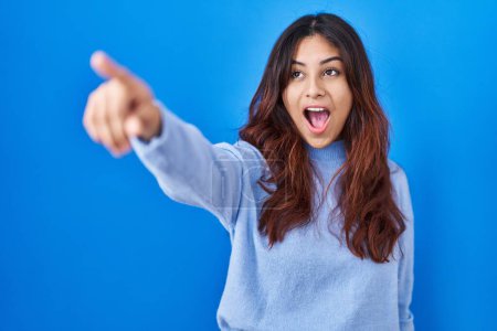 Téléchargez les photos : Jeune femme hispanique debout sur fond bleu pointant du doigt surpris devant, bouche ouverte expression étonnée, quelque chose sur le devant - en image libre de droit