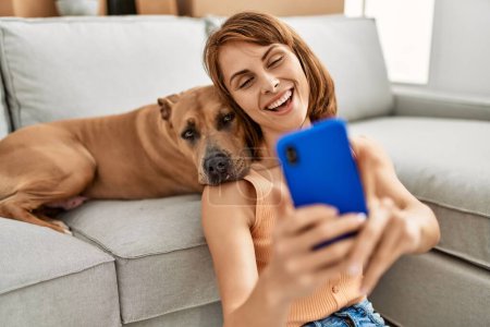 Téléchargez les photos : Jeune femme caucasienne faire selfie par le smartphone assis sur le sol avec chien à la maison - en image libre de droit